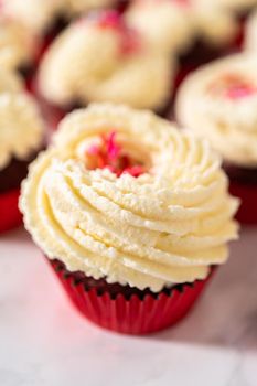 Freshly baked velvet cupcakes with white chocolate ganache frosting decorated with sprinkles.