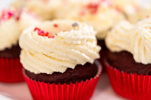 Freshly baked velvet cupcakes with white chocolate ganache frosting decorated with sprinkles.