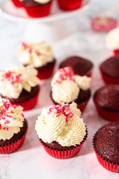 Freshly baked velvet cupcakes with white chocolate ganache frosting decorated with sprinkles.