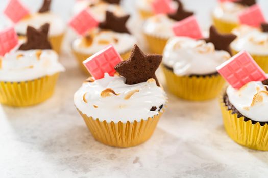 Gourmet s'mores cupcakes with meringue frosting and garnished with star-shaped chocolate graham cracker and a mini pink chocolate bar.