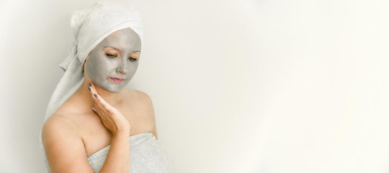 Young woman with clay cosmetic mask, wrapped in towel, touch face with hand, her gaze is directed downward. Facial skin care with cosmetic treatment. Copy space, banner