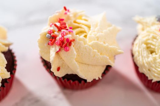 Freshly baked velvet cupcakes with white chocolate ganache frosting decorated with sprinkles.