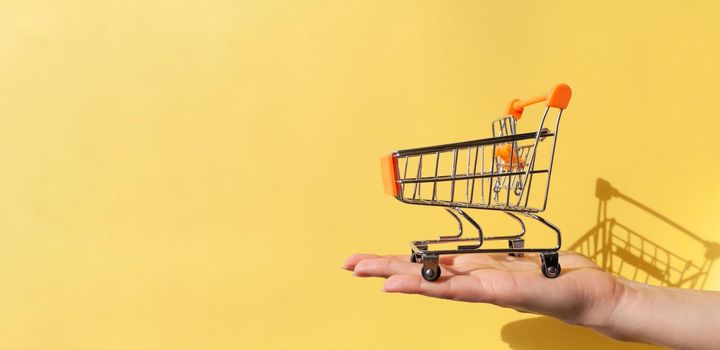 On a woman's hand is a small empty metal trolley from a supermarket. Banner. The object is on a yellow background. Horizontal photo. trading concept. Buyer. Shopping concept. Close-up. Copy space.