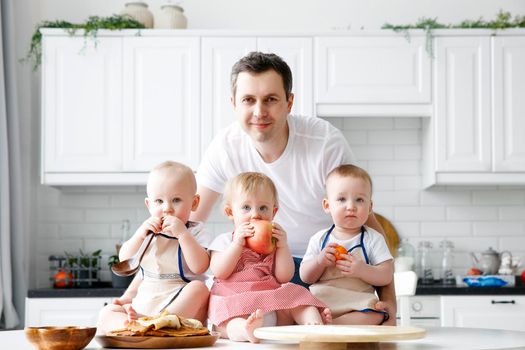 A man holds and hugs three babies in his arms. Happy father of triplets in front of a white kitchen. The concept of a happy family, eco, healthy food. Father on parental leave