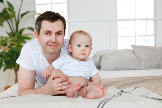 A man, a European in a white T-shirt, hugs a baby in white clothes. Bright bedroom interior on a sunny day. The concept of a young family, the rights of the child, gender equality, a happy family