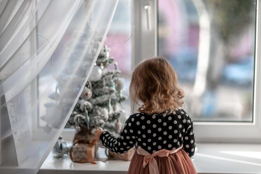 A little girl is sitting on the bed by the window and decorating a small tree with tiny Christmas toys. Happy healthy child celebrating a traditional family holiday. Adorable baby