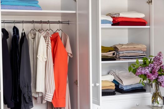 Close-up of a white wardrobe with clothes. Modern dressing room, organization of space, reasonable consumption of things. Shirts, dresses and jackets are hung on hangers. T-shirts on the shelves