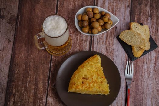 top view of typical spanish potato omelette with bread, olives, cutlery and beer mug on a wooden table with a brick wall in the background.