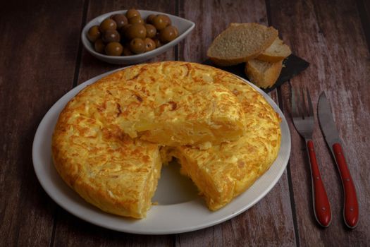 typical Spanish potato omelette with a portion cut on top accompanied by olives and sliced bread