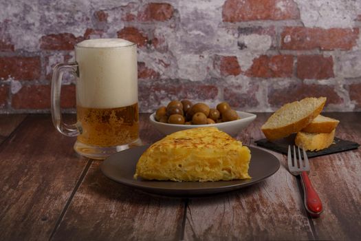 typical spanish potato omelette with bread, olives, cutlery and beer mug on a wooden table with a brick wall in the background.