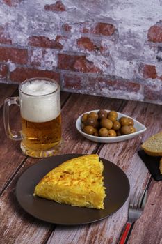 typical spanish potato omelette with bread, olives, cutlery and beer mug on a wooden table with a brick wall in the background.