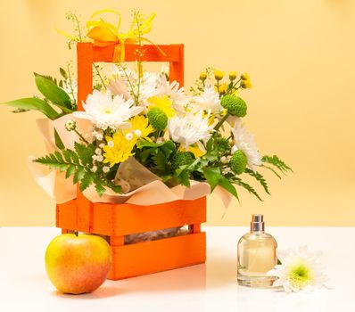 Beautiful bouquet of yellow and white flowers in the wooden basket with an apple and a bottle of perfume.