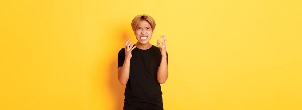 Portrait of angry and pissed-off asian blond guy, clenching hands furious and grimacing, standing over yellow background.