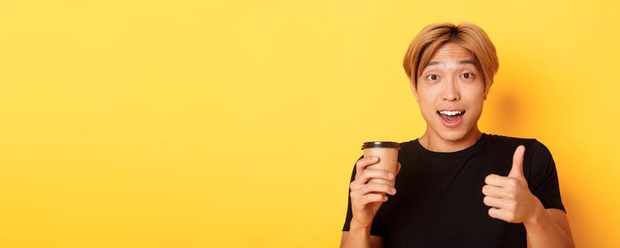 Close-up of surprised asian handsome guy recommend cafe, holding cup of coffee and showing thumbs-up in approval, smiling pleased over yellow background.