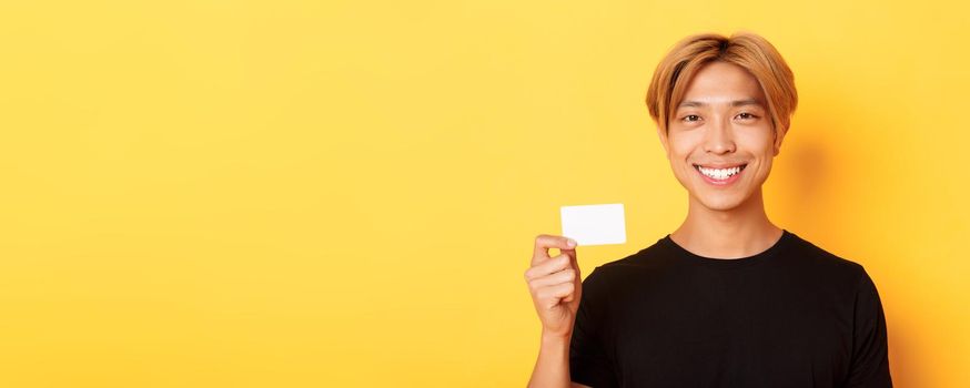 Close-up of handsome pleased asian guy with fair hair, smiling happy and showing credit card over yellow background.