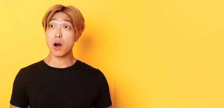 Close-up of impressed asian handsome guy, looking upper left corner with dropped jaw and amazement, standing over yellow background.