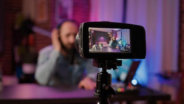 Selective focus on digital video camera screen recording male vlogger with headphones talking at microphone in home podcasting studio. Closeup of dslr filming influencer streaming live internet show.