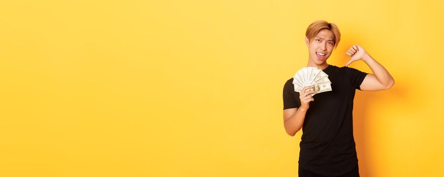 Sassy handsome asian guy pointing finger at cash and looking pleased. Korean man borrowed money, standing yellow background.