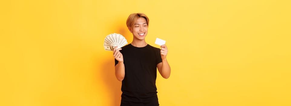 Portrait of satisfied smiling, handsome asian guy looking pleased at credit card while also holding money, standing yellow background.