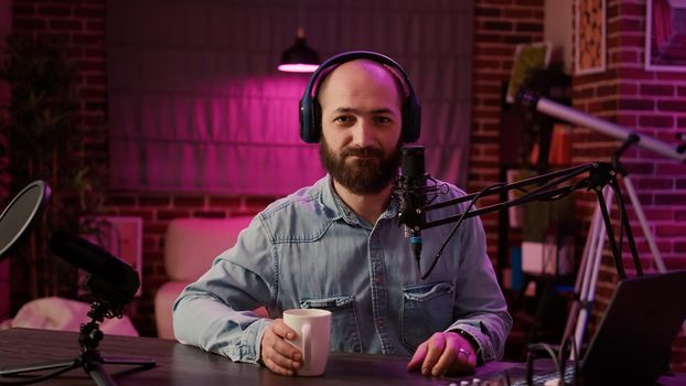 Portrait of online radio host recording podcast for entertainment show using professional audio equipment and microphone in home studio. Man influencer posing casual talking in internet broadcast.