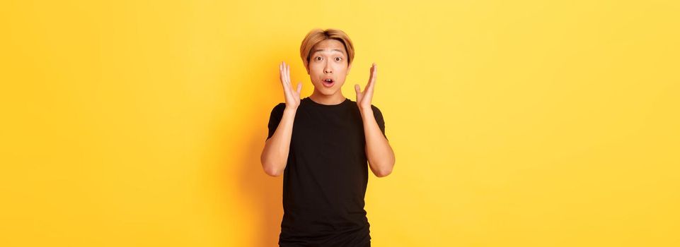 Portrait of surprised gasping blond asian guy reacting to awesome news, yellow background.