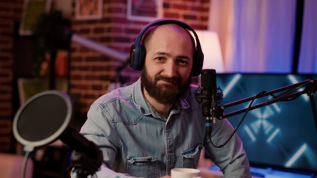 Closeup portrait of man recording online podcast with professional microphone smiling at camera sitting at desk. Online radio host with headphones streaming internet live show from home studio.