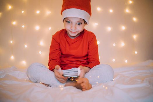 The boy is sitting in the phone on the background of a garland . New Year and Christmas. Festive mood. New Year 's attributes .