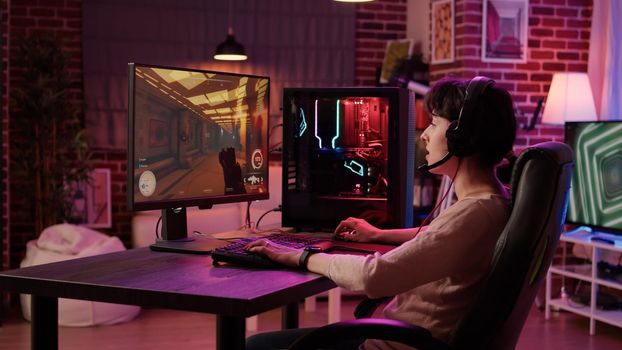 Gamer girl streaming online action game while explaining gameplay to subscribers while sitting in gaming chair. Woman using pc setup playing multiplayer first person shooter talking on headset.