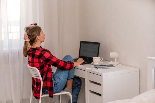 European professional woman sitting with laptop at home office desk, positive woman studying while working on PC. She is wearing a red plaid shirt and jeans