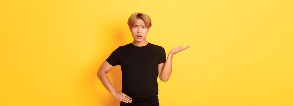 Frustrated and annoyed asian guy with blond hair, raising hand confused and looking at camera, yellow background.