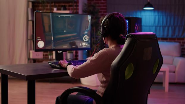 Gamer girl using pc gaming setup relaxing playing multiplayer online action game talking using headset in tournament. Woman streaming first person shooter while explaining gameplay to subscribers.