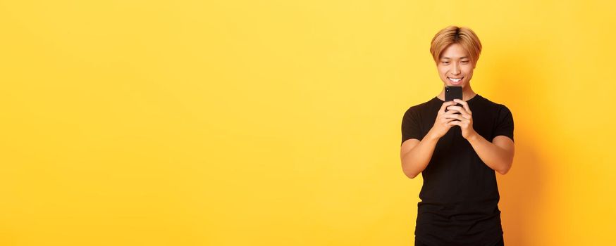 Portrait of handsome stylish asian guy with blond hair, using mobile phone and smiling, messaging in smartphone app, yellow background.