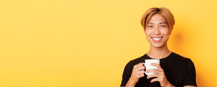 Close-up of happy satisfied asian smiling guy, holding mug with coffee, drinking and looking pleased, standing over yellow background.