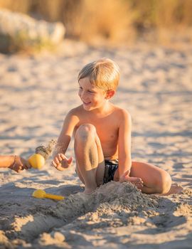 Little blond boy playing with small colorful plastic sand toy on the beach, kid building a sandcastle with fun, active happy summer holiday.