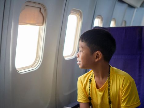 An Asian boy sits and smiles and looks out the window of an airplane.