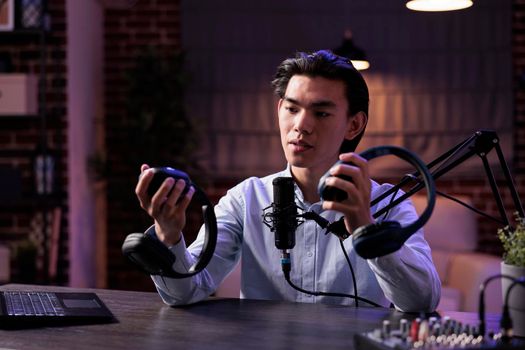 Male freelancer filming product review recommendation, recording podcast video ad talking about audio headphones on camera. Broadcasting online vlog recommending headset to channel audience.