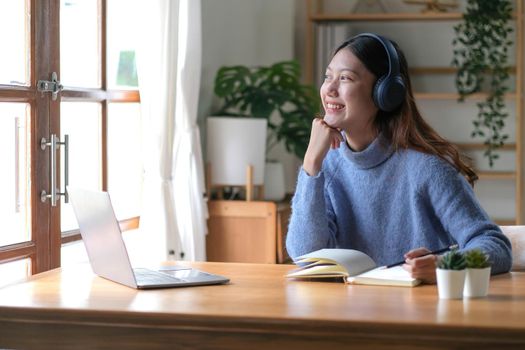 Smiling girl in headphones sitting at a desk looking outside and using a laptop to study online. Smart young women who are happy in headphones take courses on the web or practice using computers. distance education concept.