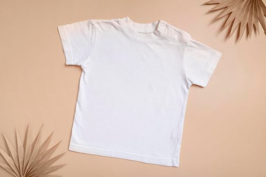 White baby t-shirt top view. Mock-up for logo, text or design on beige background. Flat lay child clothes with palm leaves.