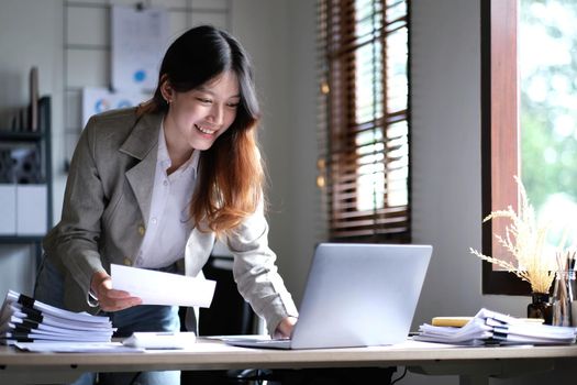 Beautiful Asian businesswoman standing and working on laptop with smiling face while working in office..