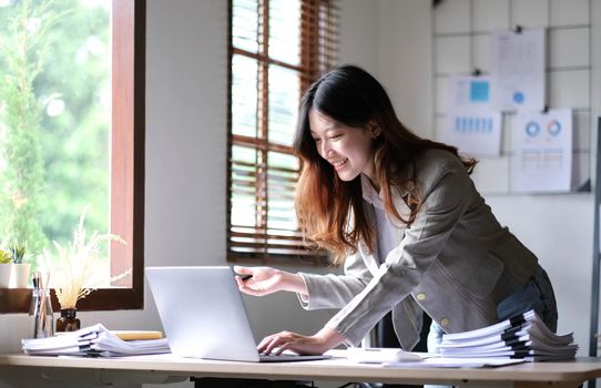 Beautiful Asian businesswoman standing and working on laptop with smiling face while working in office..