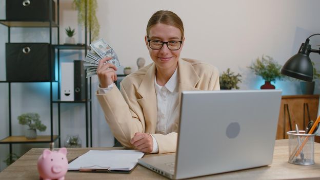 Happy businesswoman working on laptop computer sincerely rejoicing win, receiving money dollar cash banknotes, success lottery luck at office workplace. Professional manager freelancer woman dancing