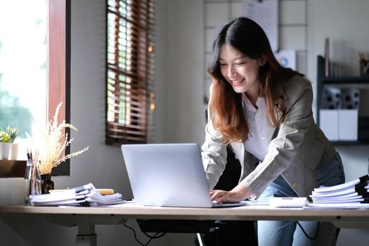 Beautiful Asian businesswoman standing and working on laptop with smiling face while working in office..