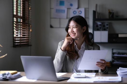 Portrait of young beautiful asian woman holding stylus pen, thinking and looking outside while sitting at office desk..