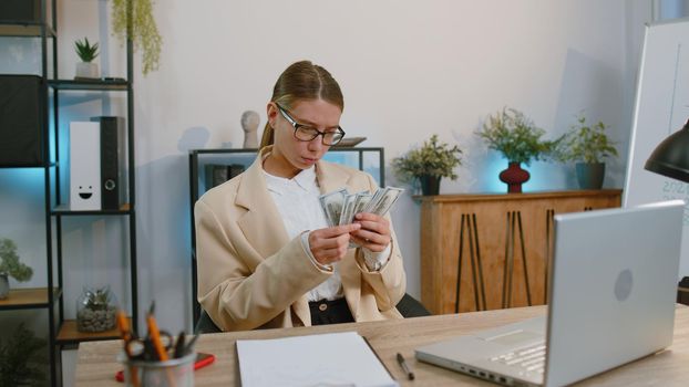 Rich happy businesswoman in suit counting money cash, calculate financial bills use laptop computer working at office workplace desk. Freelance woman satisfied of income earnings, profit. Girl worker