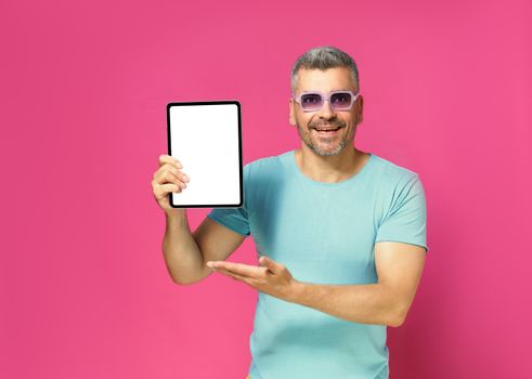 Introducing great offer showing digital tablet white screen handsome man looking at camera wearing casual blue shirt and sunglasses isolated on pink background. App advertisement.