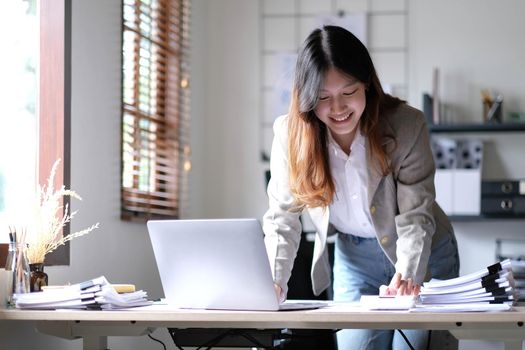 Beautiful Asian businesswoman standing and working on laptop with smiling face while working in office..