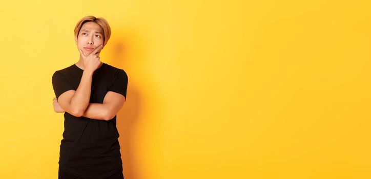 Portrait of thoughtful attractive guy in black t-shirt looking upper left corner. Young man pondering idea, standing yellow background.