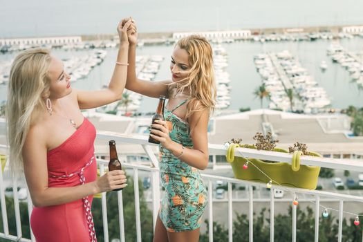 carefree young girls with beers dancing and having fun at a private party on the outdoor terrace in front of the port, leisure happiness and friendship concept