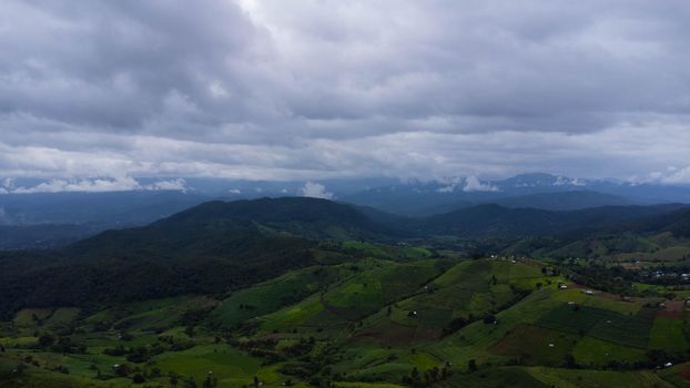 Aerial view of green wave fields with mountains and cloudy sky in rainy season. Beautiful green area of young rice fields or agricultural land in northern Thailand. Natural landscape background.