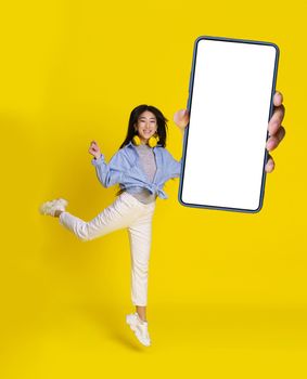 Mobile app great offer, jumping asian girl with big smartphone in hand in casual, isolated on yellow background. Full length portrait of joyful asian girl. Product placement.